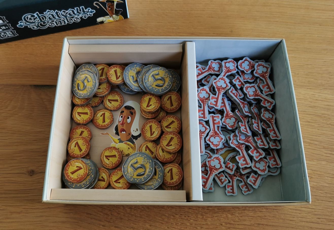 Le Fond de la boîte sert de réserve pour les pièces et les clés pendant le partie. On notera la bonne idée d'avoir placé l’illustration de la mendiante pour recevoir les pièces.